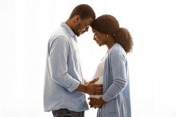 Fototapeta premium Sensual pregnant african american couple cuddling while spending time together at home, white background, copy space. Happy expecting black couple standing next to window and hugging woman big belly