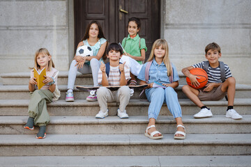 Fototapeta premium Students sitting on school steps with sport and study items showing thumbs up