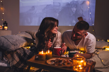 Cozy romantic evening, home weekend In bed for a loving couple. Candles, movies, hot chocolate with marshmallow, popcorn. Beautiful young man and woman enjoying relationship, having fun, laughing