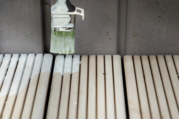 A paintbrush with white paint hovers over wooden slats, some painted and others unpainted, with a gray wooden surface visible in the background.