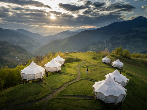 Aerial view of white domed tents perch on a vibrant green hillside, nestled beneath rolling mountains under a sky streaked with dramatic clouds, Tago, Adjara, Georgia.