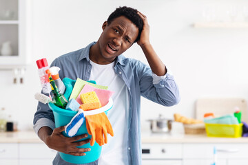 Confused black guy holding busket with cleaning tools and touching his head, unwilling cleaning house, kitchen interior. Frustrated african american man house-keeping apartment, copy space