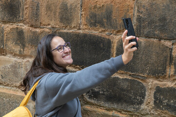Female tourist woman with yellow backpack smiling and taking a selfie with smartphone in front of an old stone wall in Edinburgh, smiling and enjoying her vacation