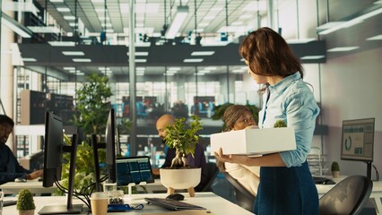 Upset worker in office packing desk plants on last day at work after quitting, saying goodbye to colleague. Employee getting fired from job during layoffs, saddening coworkers, camera B