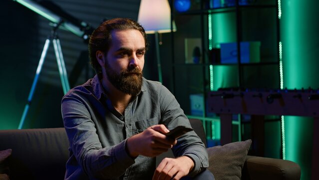 Bored person on couch in front of TV binging series on subscription based streaming services, browsing using remote. Weary man watching VOD shows on television set, zapping though channels, camera A
