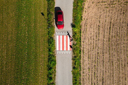 Aerial view of a red car pauses on a vibrant zebra crossing, bisecting fields of emerald and gold under the watchful lens, Poland.