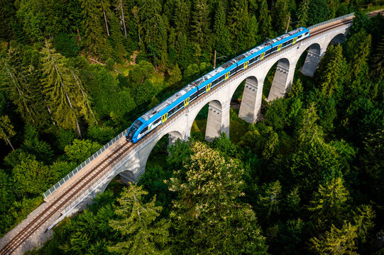 Aerial view of a bright blue and yellow train crossing a majestic stone arch bridge, cutting through a dense forest canopy, Poland.