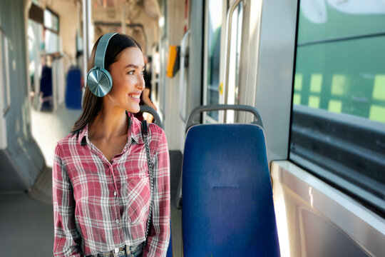 Positive European woman with headphones moving in modern city tram, female passenger enjoying trip at the public transport, looking at window and smiling - Powered by Adobe