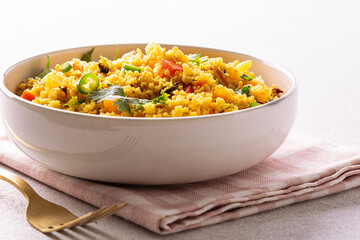 Healthy homemade vegan couscous salad with cilantro, colorful bell peppers pieces, green chilli pepper and spices in ceramic bowl on light stone table close up.