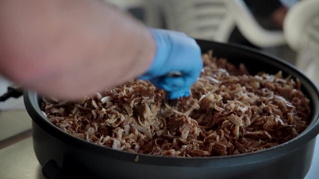 A close-up shot of a chef's gloved hands meticulously preparing and mixing freshly cooked pulled pork or shawarma meat in a large black pot with a fork