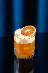 Glass of amber-colored cocktail with foam and a dried orange slice on top, set on a glossy black surface with a dark blue background