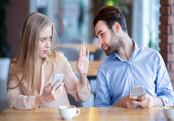 Young guy trying to look into his girlfriend's phone at cafe, feeling jealous, young lady stopping him. Smartphone and social media addiction, phubbing, relationship problems concept