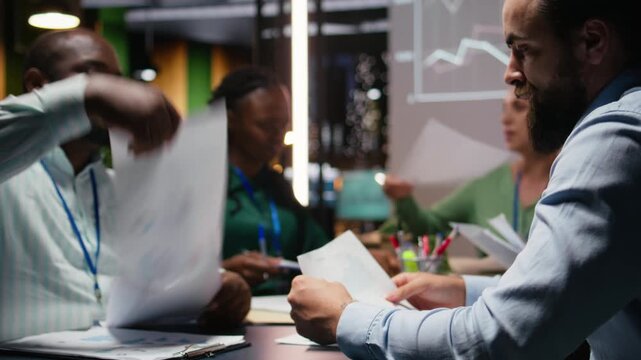 Diverse project team shares analytics reports during advisory board event after hours, reviewing business intel and preparing insight for upcoming presentation with advisors. Camera A.