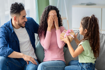 Portrait of cute little girl surprising her arab mom, giving present at home, daughter greeting young mum with Mother's day or birthday, excited lady covering eyes while sitting on couch with husband