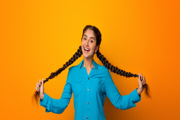 Portrait of smiling happy young indian woman holding and pulling her two long braids, playing with hair, looking at camera, cheerful lady posing in studio, isolated over yellow background, free space