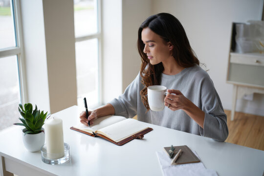 Thoughtful woman journaling with morning coffee at cozy home office desk for lifestyle blog