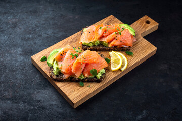 Pumpernickel bread sandwich snack with cured smoked salmon and fresh avocado served as close-up on a wooden cutting board