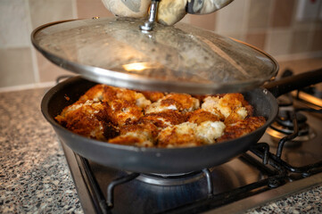 A person wearing an oven mitt lifting the lid off a frying pan full of breaded chicken, revealing the cooking process on a home stove. The scene captures the moment of checking the meal while cooking