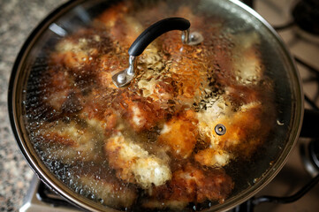 Close-up of breaded chicken cooking in hot oil, covered by a transparent lid. Steam and bubbling oil create an authentic, in-progress cooking shot. High quality photo