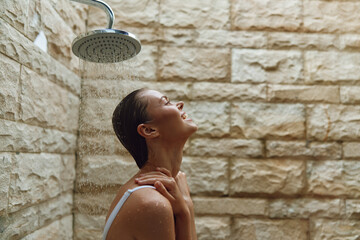 smiling woman enjoying outdoor shower with stone wall background, fresh water drops, relaxed mood, natural light, casual white swimsuit