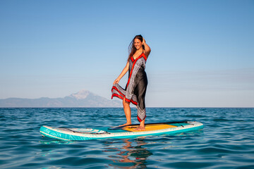 Young attractive woman in dress standing on sup board in water. Happy pretty  woman relaxing on a...