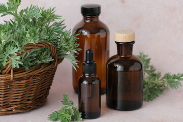 Brown glass bottles filled with essential oils are placed on a soft backdrop, accompanied by a small basket of fresh herbs. Alternative medicine, healthy wormwood and well-being concept. 