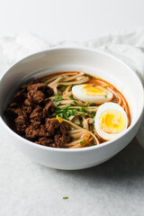 Overhead view of spicy noodle soup with eggs and beef chunks in a white bowl, top view of noodles eggs in a spicy broth with scallions