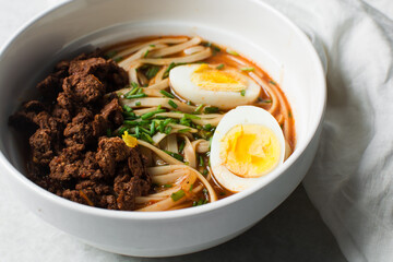 Overhead view of spicy noodle soup with eggs and beef chunks in a white bowl, top view of noodles eggs in a spicy broth with scallions