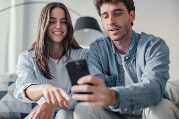 Happy millennial couple sit relax on couch in living room watching video on cellphone together,...