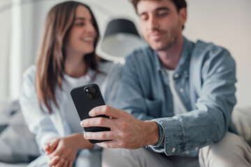 Happy millennial couple sit relax on couch in living room watching video on cellphone together,...