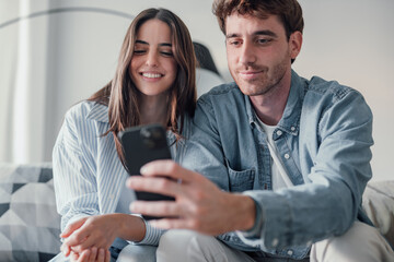 Happy millennial couple sit relax on couch in living room watching video on cellphone together,...