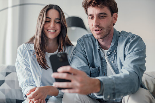 Happy millennial couple sit relax on couch in living room watching video on cellphone together, smiling young husband and wife rest on sofa at home browsing Internet using modern phone device