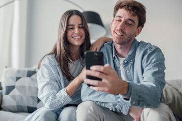 Fototapeta premium Cute couple greeting having fun video calling long distance friends and family catching up. Young man holding phone with girlfriend sitting on the sofa laughing and enjoying technology