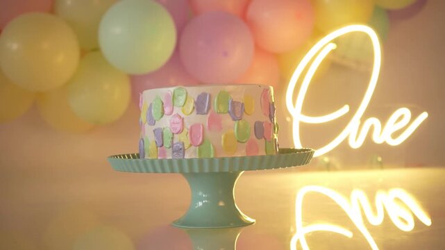 Horizontal panning shot of a first birthday smash cake setup, featuring a pastel-toned cake, a neon 'ONE' sign, and a balloon garland, all arranged in a beautifully decorated scene 