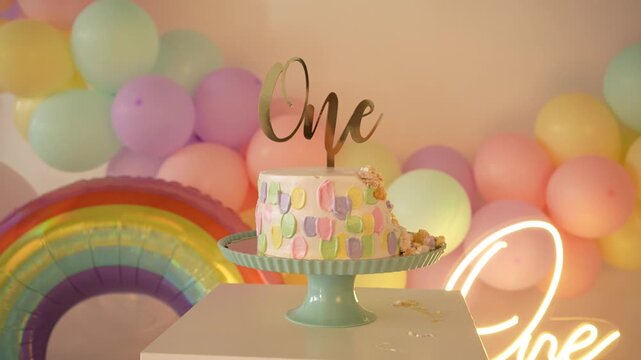 Left-to-right horizontal pan of a 1st birthday smash cake setup with bitten pastel cake in close-up, ONE topper, neon ONE sign, balloon garland, and a rainbow balloon in frame