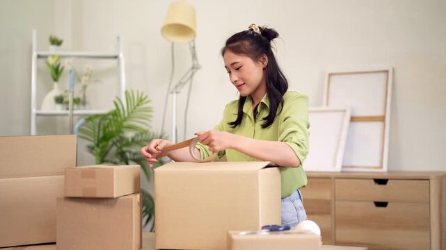 Asian adult woman packs cardboard box sealing personal items for house move relocation shipping parcels organizing new home preparations home office
