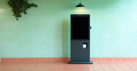 Kiosk stands against pale green wall. Red tile floor, plant visible above. Simple lighting fixture present. Minimalist design.