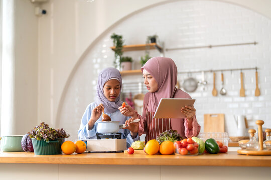 Muslim girl cooking with woman using tablet for recipe, halal food learning with technology, warm islamic family lifestyle, digital nutrition education for muslim kid, modern halal kitchen inspiration - Powered by Adobe