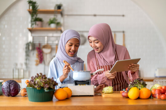 Muslim girl cooking with woman using tablet for recipe, halal food learning with technology, warm islamic family lifestyle, digital nutrition education for muslim kid, modern halal kitchen inspiration