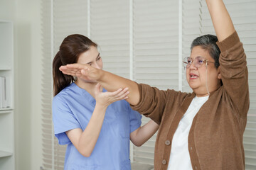 Fototapeta premium Senior asian woman lifts one arm in upward diagonal motion assisted by asian female physiotherapist during shoulder rehabilitation session focused on strength and mobility recovery
