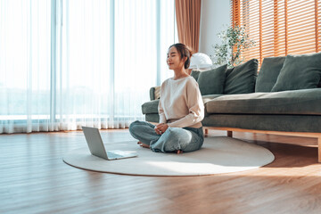 Happy young Asian woman practicing yoga and meditation at home sitting on floor in living room in lotus position with laptop