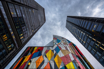 Low-angle shot of two modern skyscrapers, one featuring a vibrant, colorful, geometric facade, juxtaposed against a darker, more traditional skyscraper.