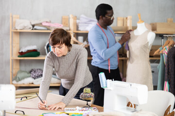In workshop, female tailor designer draws diagram of future clothing product on paper. Creating dress pattern according to individual sizes.African companion work in background with mannequin