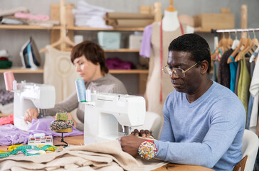 Woman designer and African male apprentice sews clothes using sewing machine. Worker creates wardrobe items, sews dresses and suits for clients, and makes clothes.