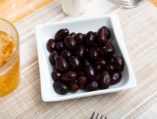 On small square white plate lies portion of large ripe black olives with stone.Snack is supplemented with glass of foamy beer