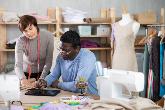 Two interested fashion designers, man and woman, collaborating in sewing studio, using tablet to create and refine new garment design at pattern table, discussing digital sketches or client references