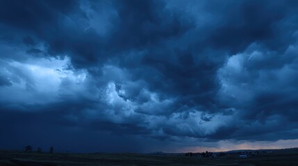 Fototapeta premium Dark storm clouds loom ominously over a vast countryside, creating a dramatic atmosphere during the twilight hours. The sky transitions to blue as the light fades.