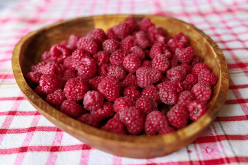 A wooden bowl with ripe raspberries lies on a rustic tablecloth.