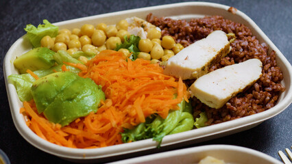 Balanced and nutritious lunch box presents grilled chicken breast, brown rice, chickpeas, avocado slices, shredded carrots, and lettuce, promoting a healthy and wholesome meal