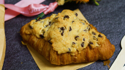 Freshly baked crookie croissant filled with a chocolate chip cookie, resting on baking paper, with a pink ribbon and dried flowers in the background, creating a tempting pastry scene